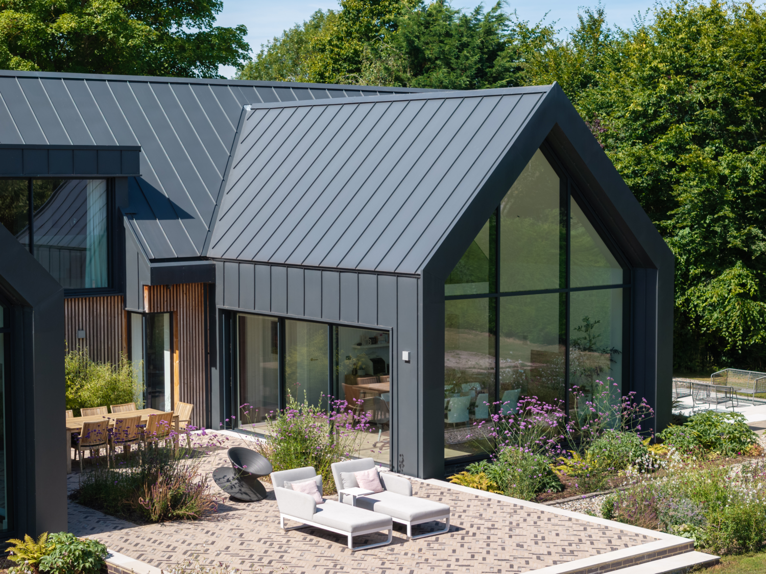 Modern house with large glass windows and a sleek, dark metal Catnic Urban roof and cladding. Patio features white lounge chairs and a dining table. Surrounded by lush greenery and vibrant garden plants.