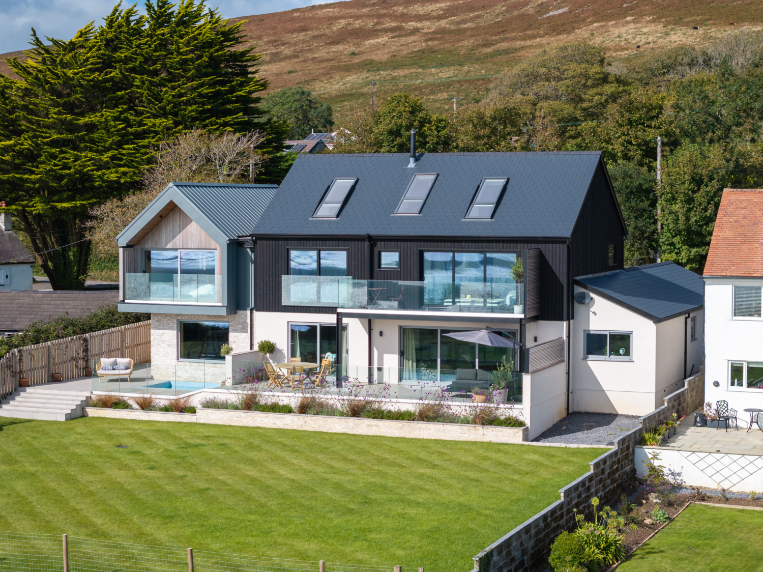 Modern house with large glass windows and dark metal Catnic Urban cladding and roofing, surrounded by lush greenery. The patio features outdoor seating, including a dining table and lounge chairs, amid landscaped gardens.